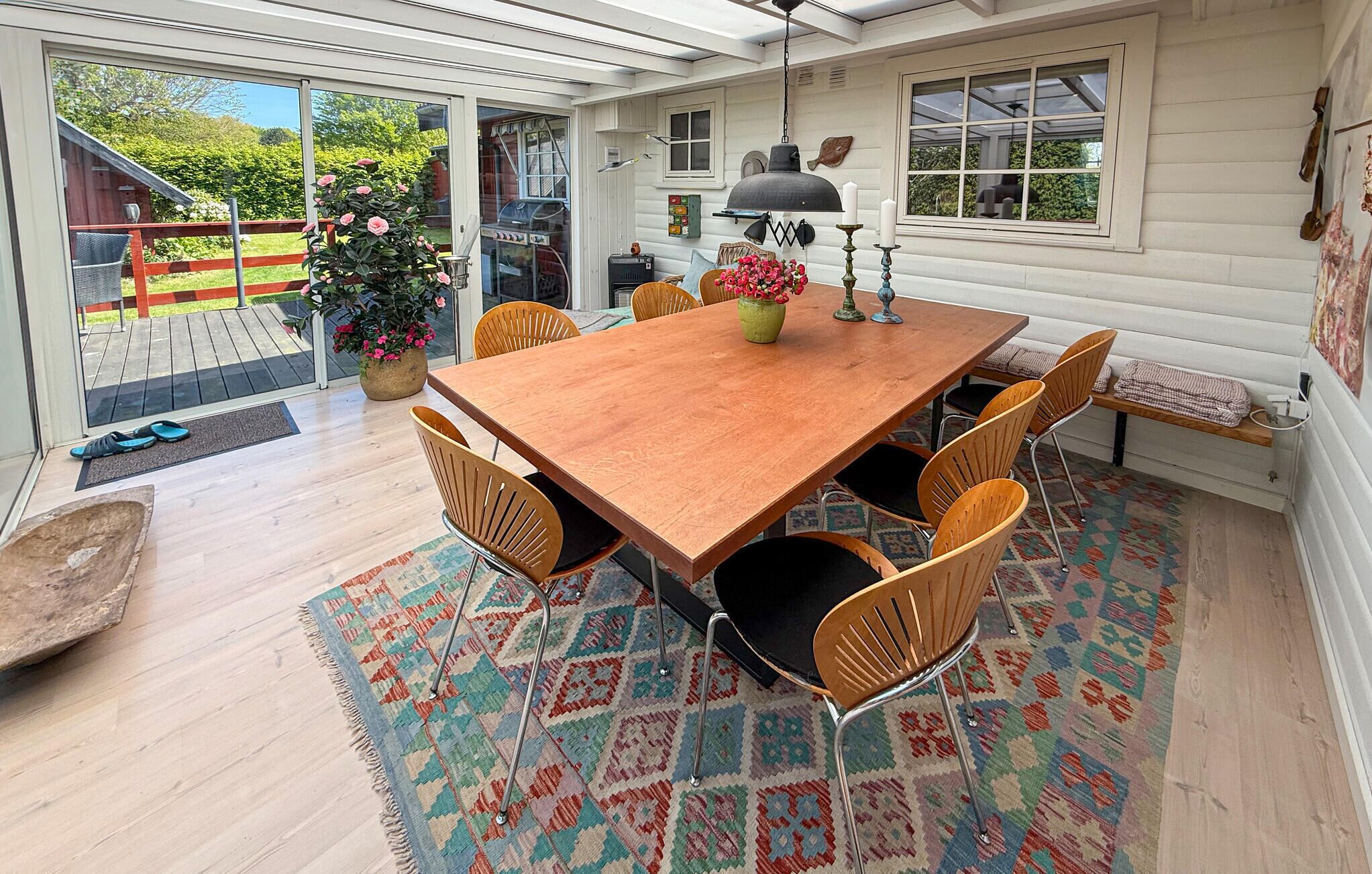 A wooden dining table with six chairs, a patterned rug, and a potted plant with pink flowers.