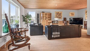 En rummelig stue med træloft, et stort vindue, et træskab, en sofa, et fjernsyn og en reol.