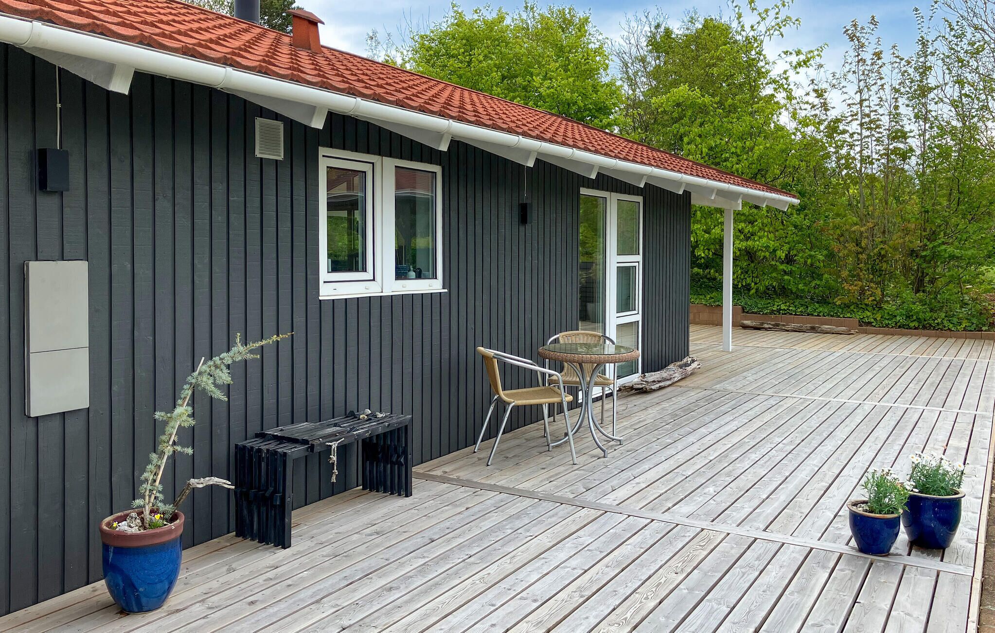 Ein graues Gebäude mit roten Ziegeln auf dem Dach, einer Holzterrasse und einer Glastür, die zu einem Außenbereich mit Sitzgelegenheiten führt.