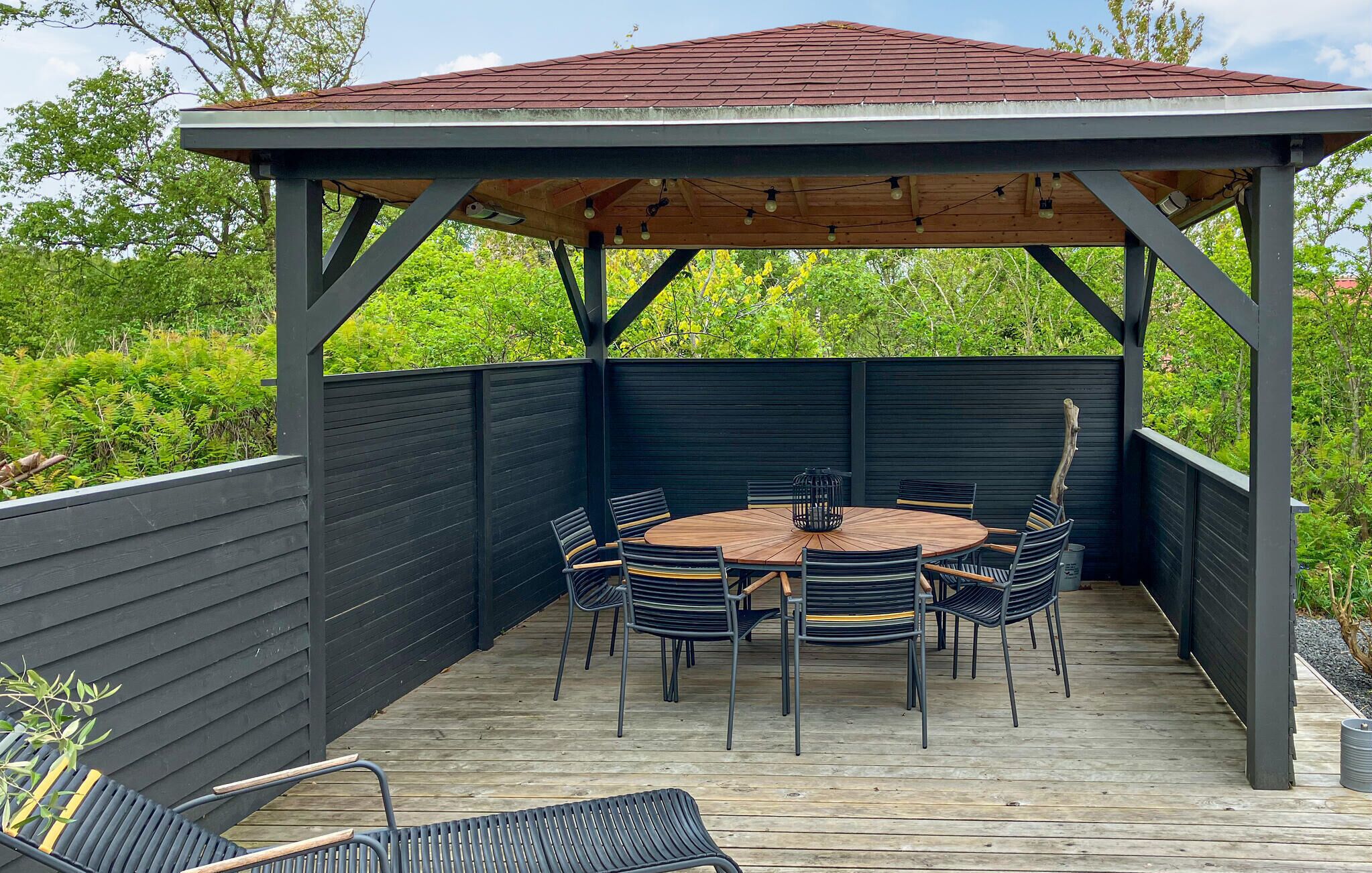 A covered outdoor patio with a round table and chairs, surrounded by a wooden deck and a black fence.