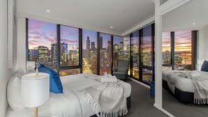 A modern hotel room with a large bed, a chair, and a view of a city skyline at dusk. - Exclusive Stays - Vue Grande (Southbank)