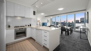 A modern kitchen with white cabinets, a gas stove, and an oven. There is a dining area with a table and chairs, and large windows offering a city view. - Exclusive Stays - Vue Grande (Southbank)