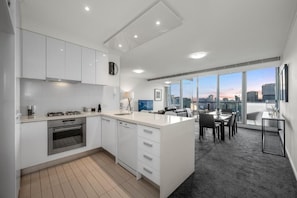 A modern kitchen with white cabinets, a gas stove, and an oven. There is a dining area with a table and chairs, and large windows offering a city view. - Exclusive Stays - Vue Grande (Southbank)