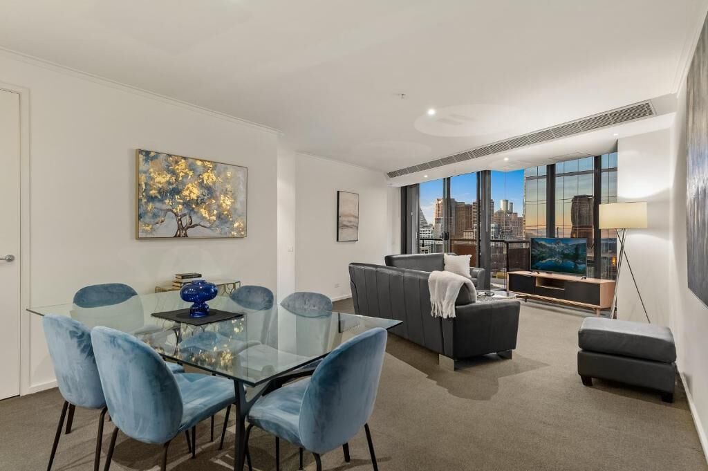 A modern living room with a glass dining table, blue chairs, a grey sofa, and a city view through large windows.