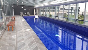 An indoor swimming pool with a glass ceiling, benches, and a view of the cityscape. - Exclusive Stays - Vue Grande (Southbank)