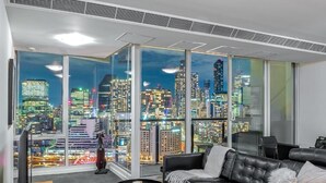 A modern living room with a cityscape view, a leather sofa, a small table, and a large window. - Exclusive Stays - Vue Grande (Southbank)