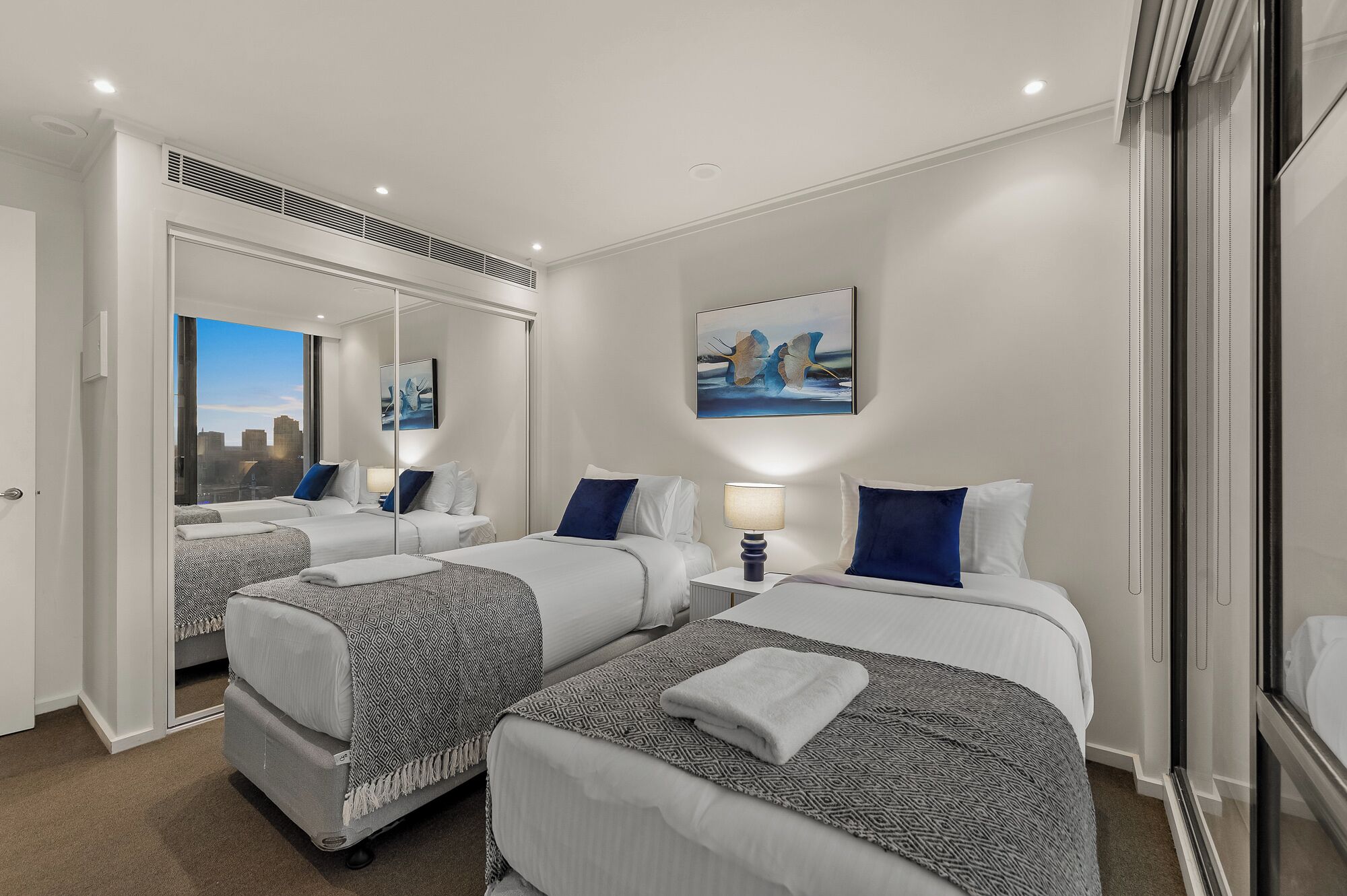A hotel room with two beds, a large mirror, a painting, and a city view.