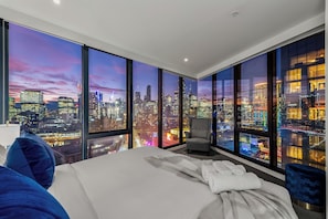 Une chambre d’hôtel moderne avec un grand lit, une chaise et une vue sur le paysage urbain illuminé la nuit.