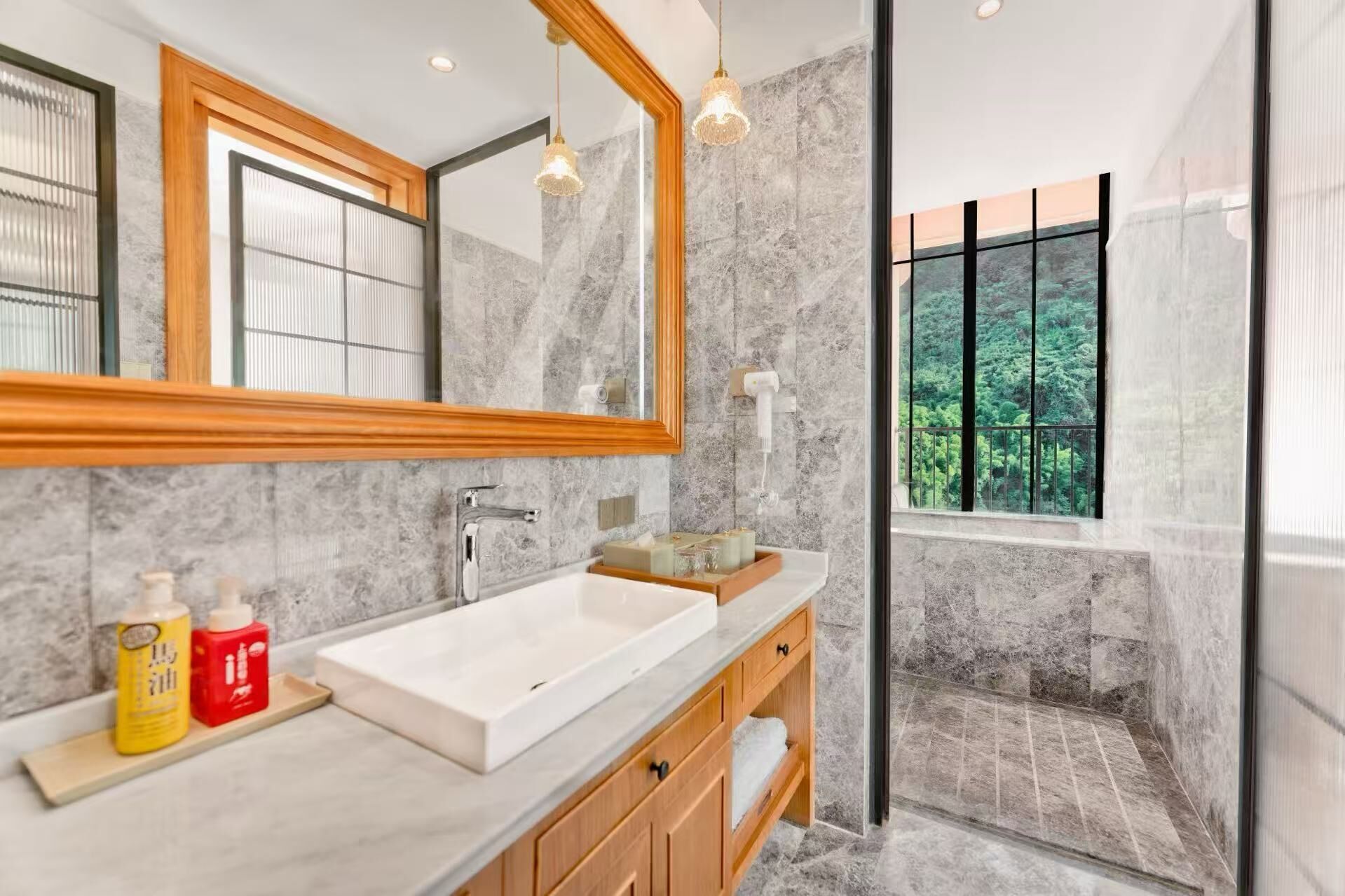 Une salle de bain moderne avec un grand miroir, un lavabo blanc, des meubles en bois et un espace douche donnant sur un paysage verdoyant.