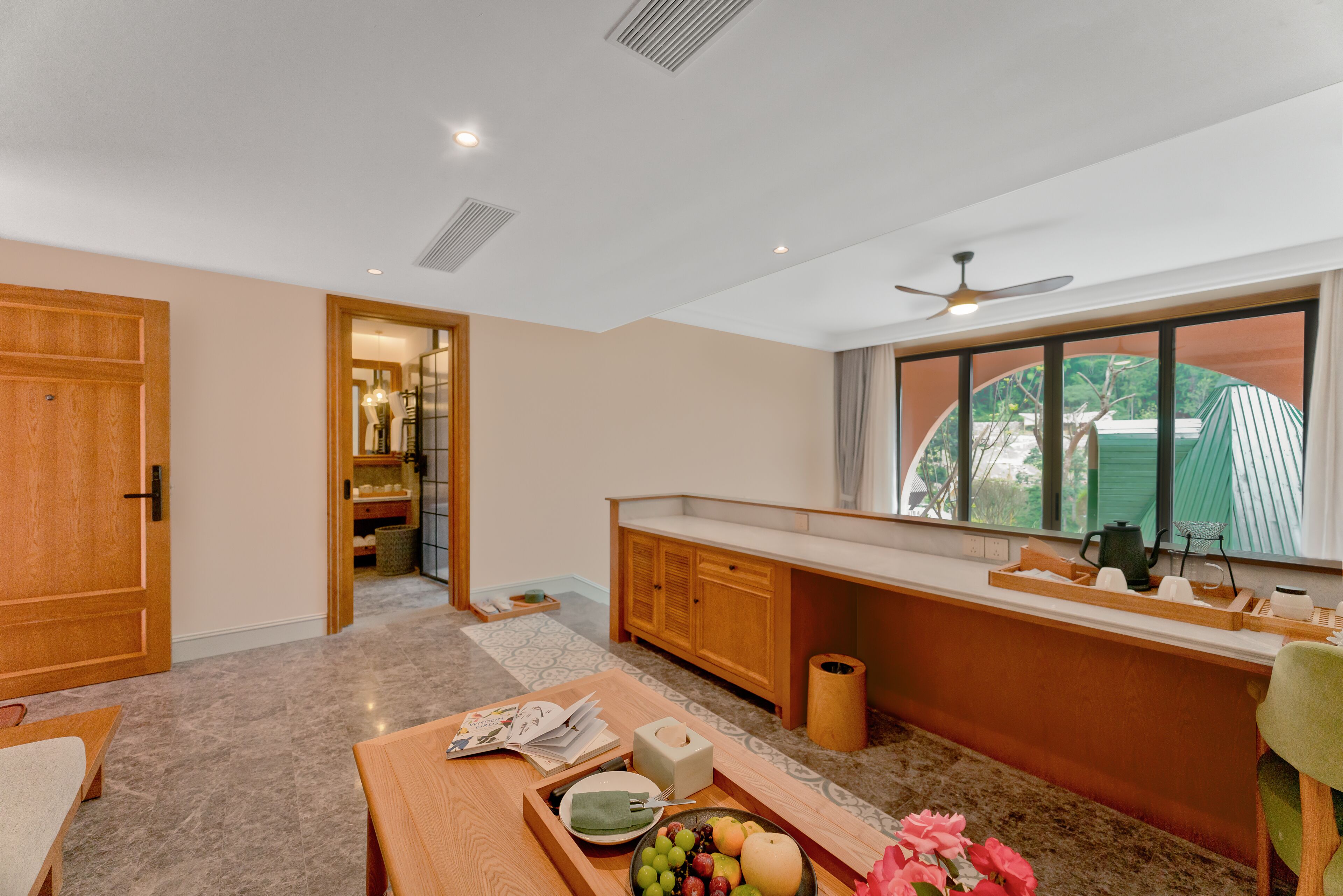 A room with a wooden cabinet, a dining table with a fruit bowl, a window with a view, and a ceiling fan.