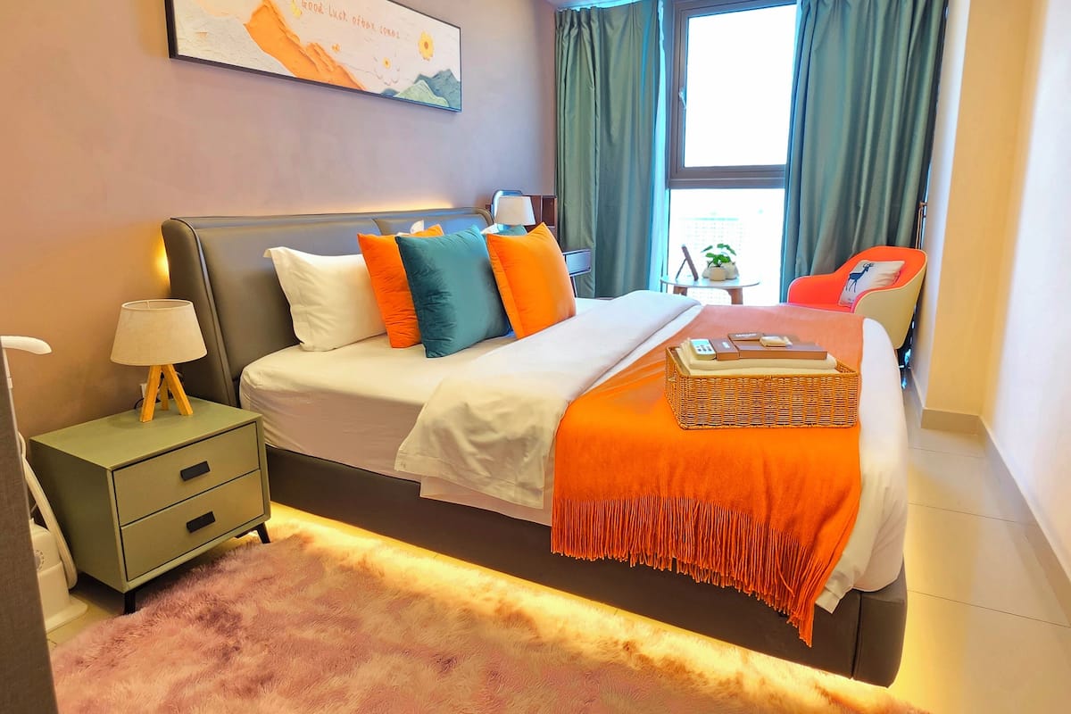 Una habitación de hotel moderna con una cama grande, una mesita de noche, una lámpara, una pequeña mesa con una planta y vistas al exterior a través de una ventana.