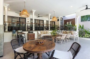 A spacious indoor area with a round wooden table, wicker chairs, and a sofa with cushions. There are ceiling fans, a water dispenser, and a view of the outside through large windows. - 3BDR Villa Baruna, Good Location (Seminyak)