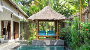 Ein Pool mit einem strohgedeckten Gazebo, einer Holzbank und üppiger Bepflanzung.