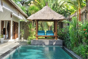 Ein Pool mit einem strohgedeckten Gazebo, einer Holzbank und üppiger Bepflanzung.