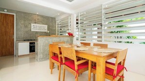 A modern dining area with a wooden table and chairs, a kitchen in the background, and large windows with blinds. - Sweet Villa Love Umalas! Breakfast included (Umalas)