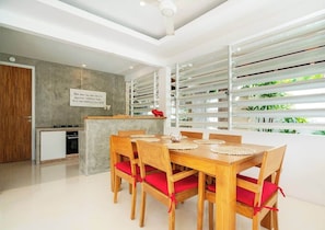 A modern dining area with a wooden table and chairs, a kitchen in the background, and large windows with blinds. - Sweet Villa Love Umalas! Breakfast included (Umalas)