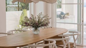 Spiseplads med et træbord, hvide stole, en vase med blomster og udsigt gennem en glasdør.