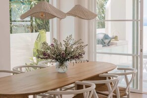 A dining area with a wooden table, white chairs, a vase with flowers, and a view through a glass door. - Villa Lola with Ocean View & Breakfast (Uluwatu)