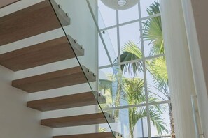 A staircase with glass railings and wooden steps, leading to a room with a large window overlooking palm trees. - Villa Lola with Ocean View & Breakfast (Uluwatu)