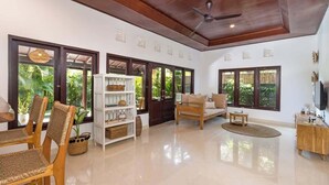 A spacious living area with a wooden ceiling, large windows, a white bookshelf, a sofa, and a TV.