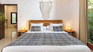 A bedroom with a large bed, two bedside tables with lamps, a macrame wall hanging, and a view of greenery through a window.