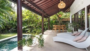A covered outdoor area with a pool, lounge chair, and dining table surrounded by lush greenery.