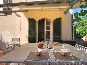 Ein Tisch, gedeckt mit herzförmigen Tassen und einem Teekännchen, umgeben von weißen Stühlen, mit Blick auf ein Gebäude mit grünen Fensterläden und einem großen Fenster im Hintergrund.