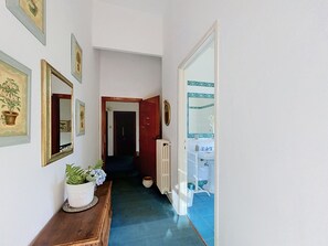 Ein Flur mit einer hölzernen Bank, Topfpflanzen, gerahmten Bildern an der Wand, einem Heizkörper und einem durch eine offene Tür einsehbaren Badezimmer.