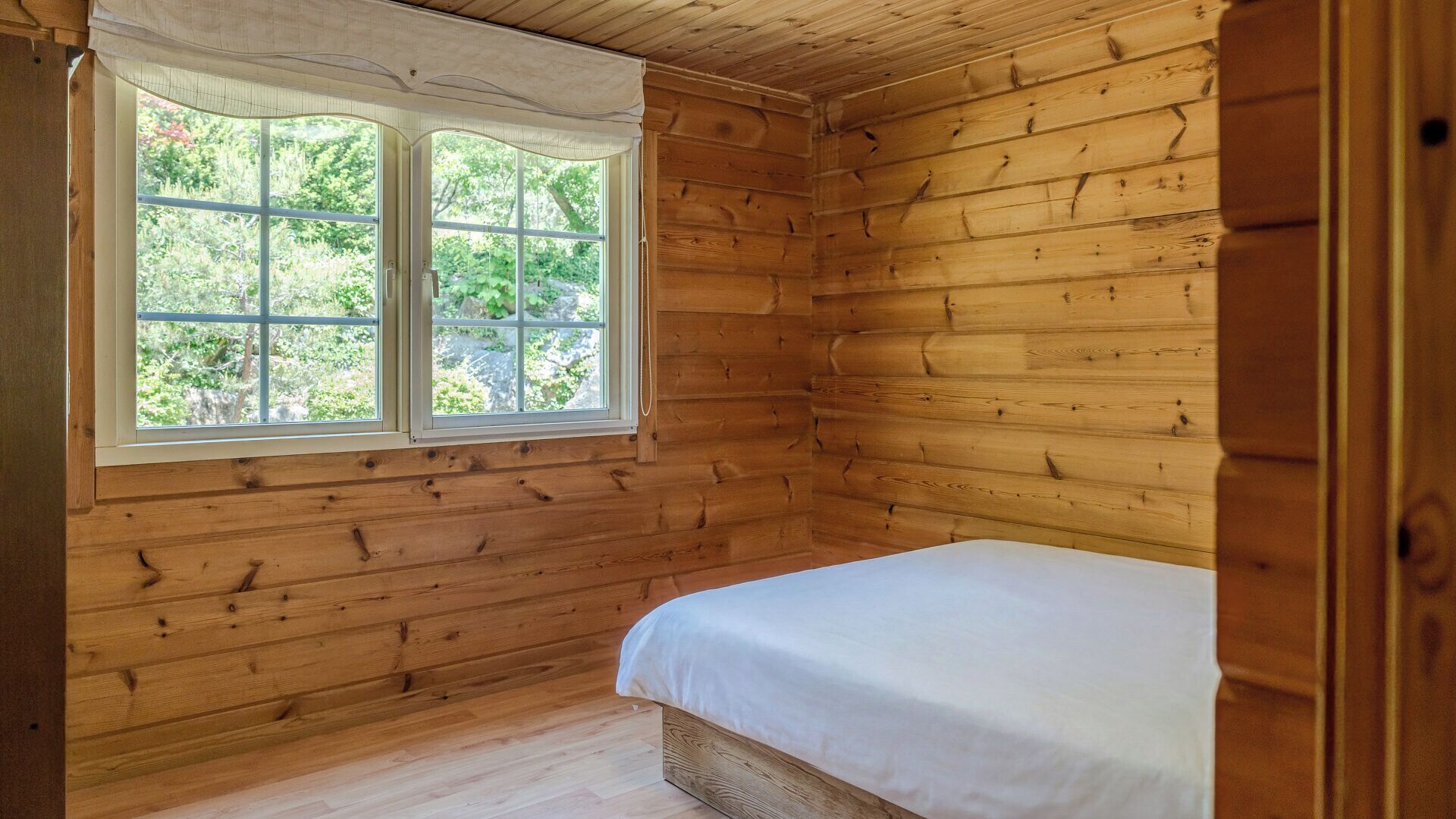 A wooden cabin room with a bed, a window with a view of greenery, and a wooden floor.