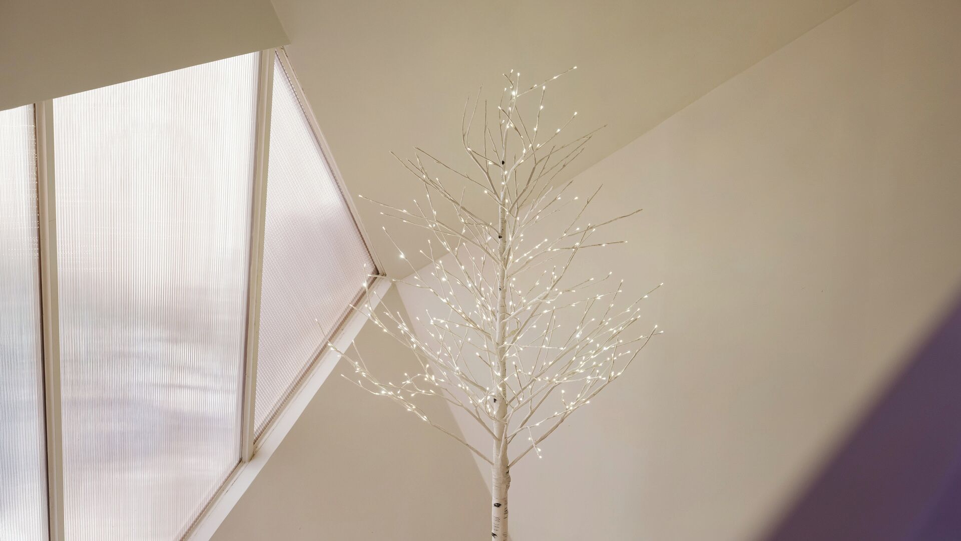 Una habitación con una estructura blanca que recuerda a un árbol, decorada con luces, una ventana con persianas y una pared lisa.