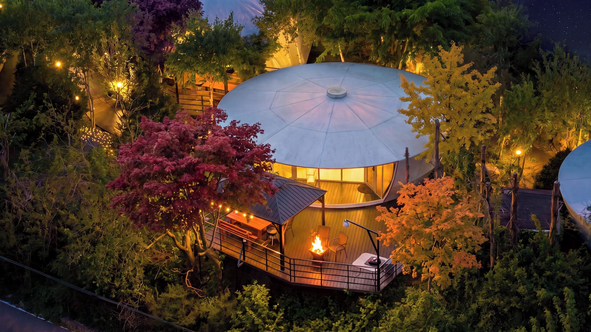 Un bâtiment circulaire surmonté d’un dôme, entouré d’arbres, doté d’une terrasse en bois et agrémenté d’un foyer.