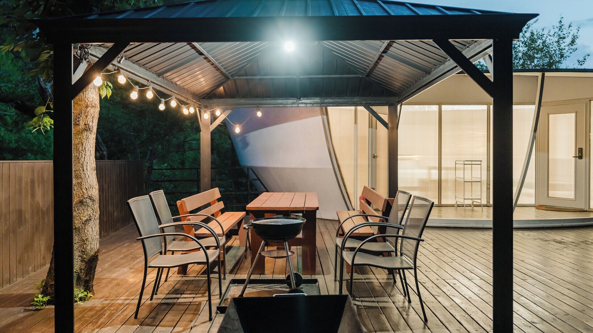 Een terras buiten met een houten tafel en stoelen, een barbecue en lichtslingers.
