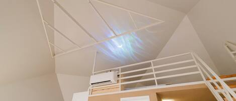 Une chambre avec un lit superposé, un projecteur au plafond diffusant un ciel étoilé et un appareil de climatisation blanc.