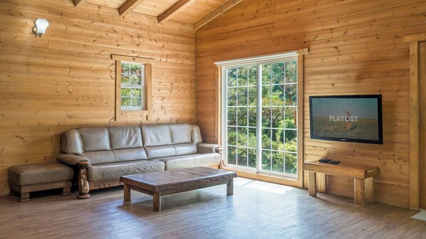 Interior de una cabaña de madera con sofá, mesa de centro, televisor y ventanas amplias.