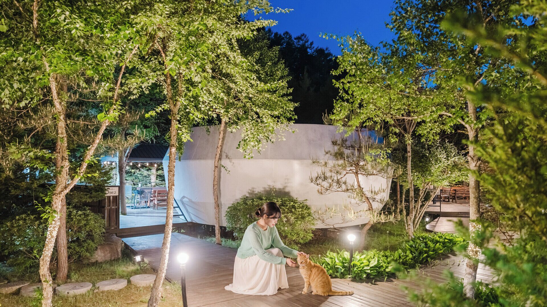 Une femme se trouve dans un jardin, avec un chat, des allées en bois et un bâtiment en arrière-plan.