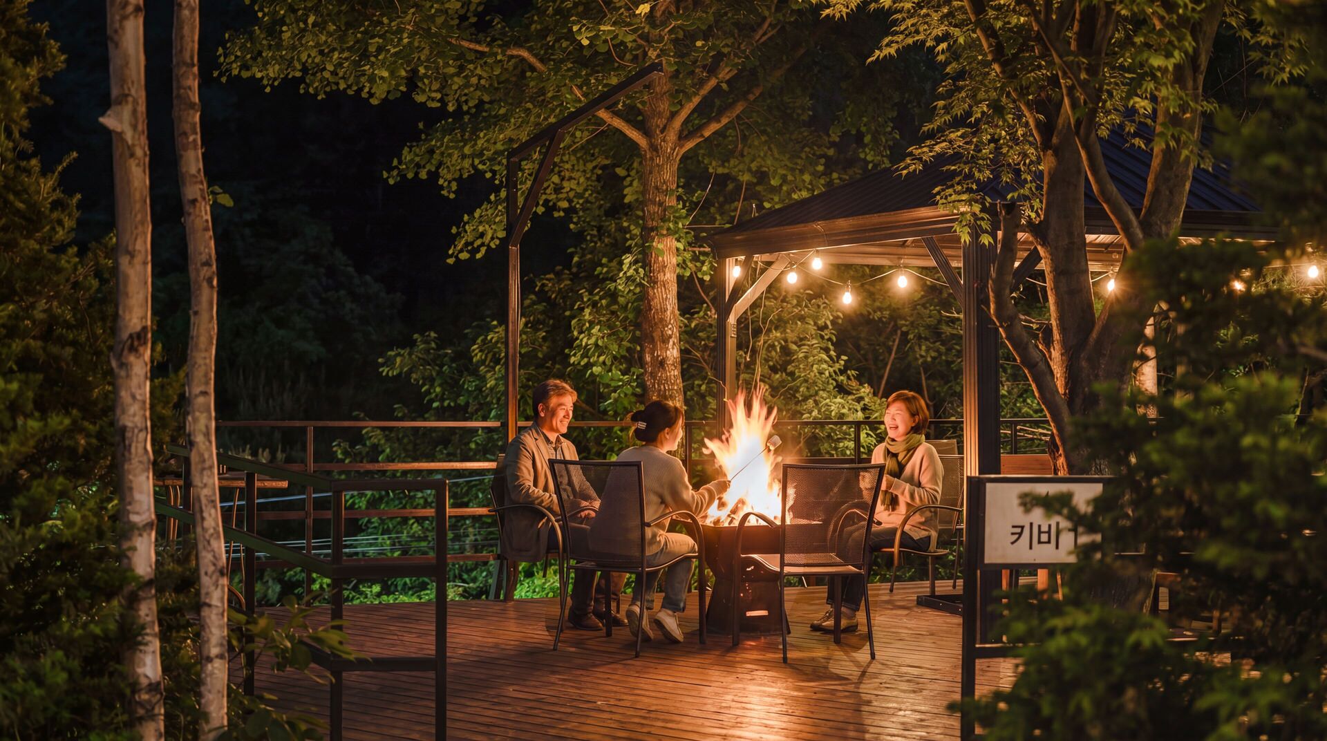 Un espace extérieur aménagé, partiellement abrité, avec un foyer, entouré d’arbres et doté d’une terrasse en bois.