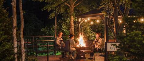Un espace extérieur aménagé, partiellement abrité, avec un foyer, entouré d’arbres et doté d’une terrasse en bois.