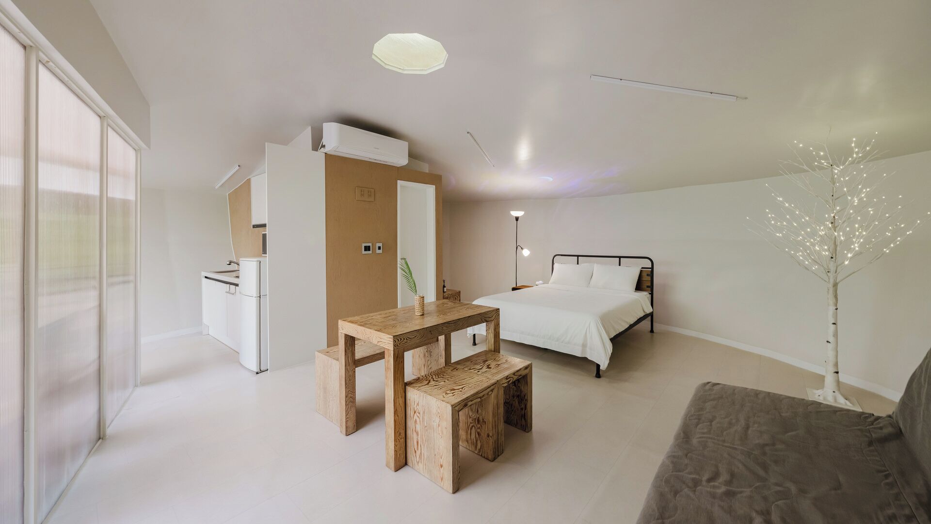 A modern hotel room with a bed, a desk, a chair, a sofa, a bedside table, and a decorative tree.