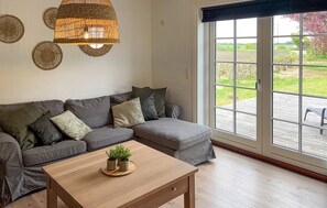 Ein Wohnzimmer mit einer grauen Eckcouch, einem Couchtisch aus Holz mit Pflanzen, einer gewebten Leuchte und einer großen Schiebeglasdoor, die auf eine Terrassenanlage führt.