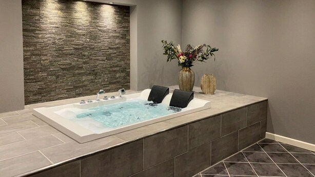 Ein modernes Badezimmer mit Steinwand, einer großzügigen Badewanne, zwei schwarzen Stühlen und einer Vase mit Blumen.