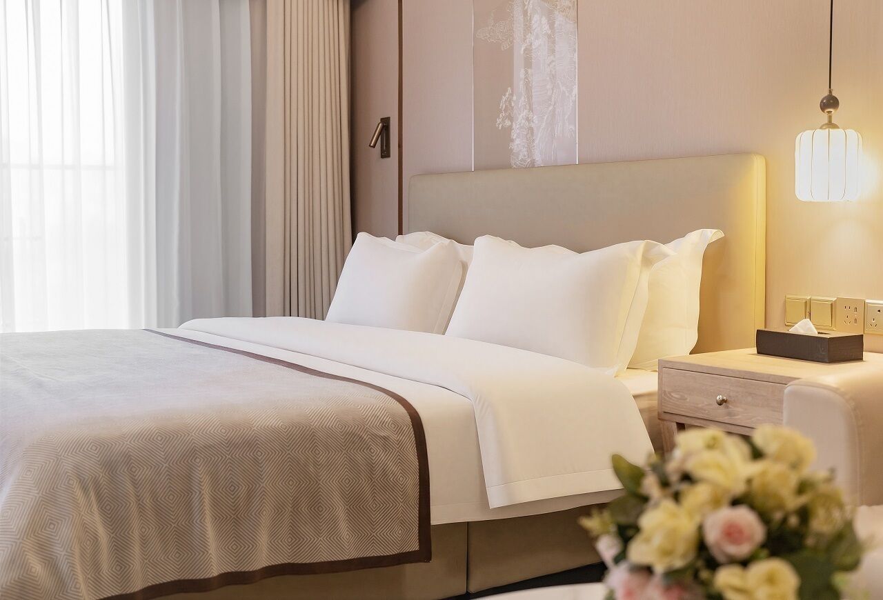 A hotel room with a bed, bedside table, lamp, and a vase of flowers.