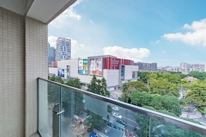 A city view from a balcony with a glass railing, a large building with advertisements, and surrounding greenery.