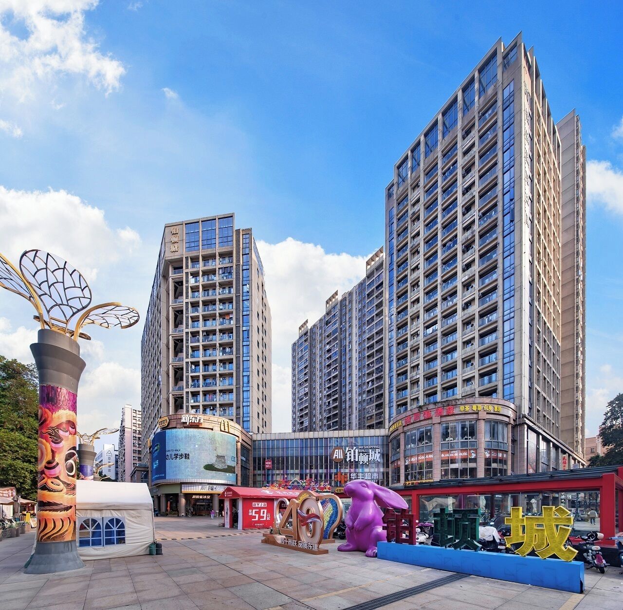 A modern urban area with high-rise buildings, a shopping center, and a prominent sculpture.