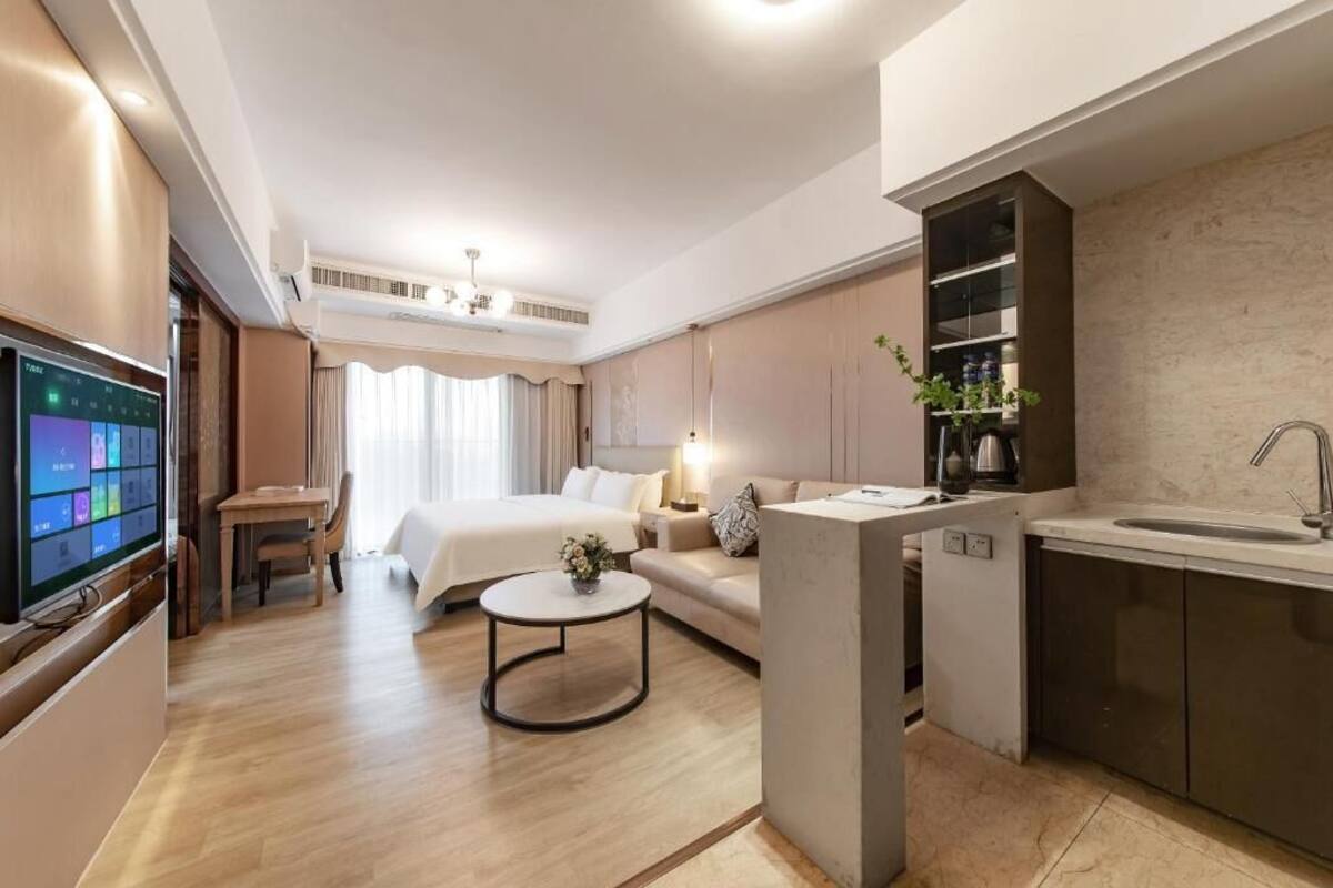 A modern hotel room with a bed, a desk, a small round table, a kitchenette, and a TV.