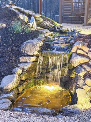 Ein Wasserfall aus Stein mit einem kleinen Teich am Fuße, umgeben von Steinen und Kieselsteinen.