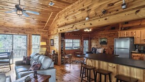 Interior de una cabaña con techo de madera, zona de comedor, cocina con refrigerador y sala de estar con sofá y sillones.