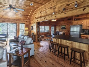 Ein Interieur einer Holzhütte mit Holzbalkendecke, einem Essbereich, einer Küche mit Kühlschrank und einer Sitzecke mit Sofa und Sesseln.