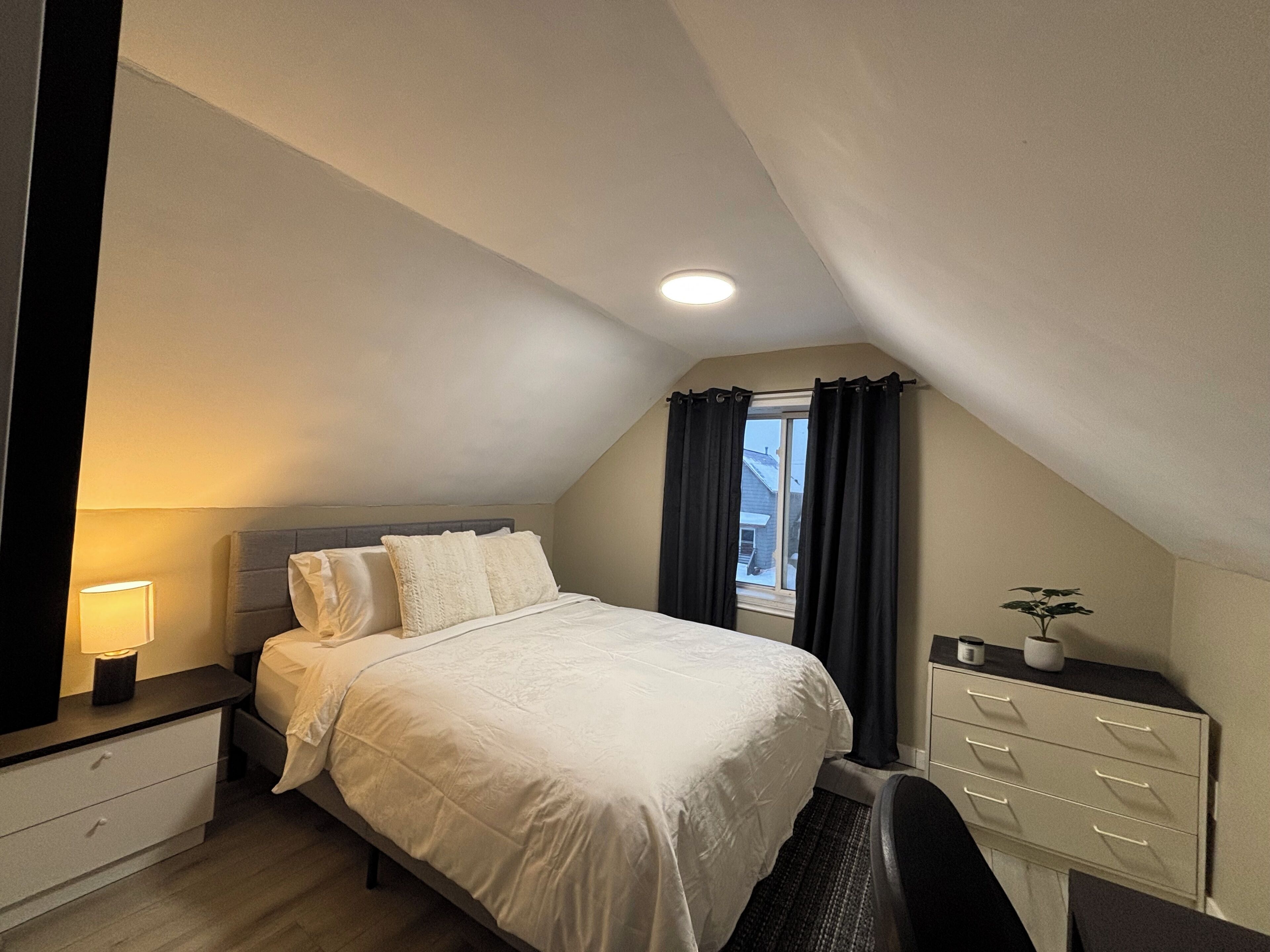 Un dormitorio con cama, mesita de noche, lámpara y ventana con cortinas.