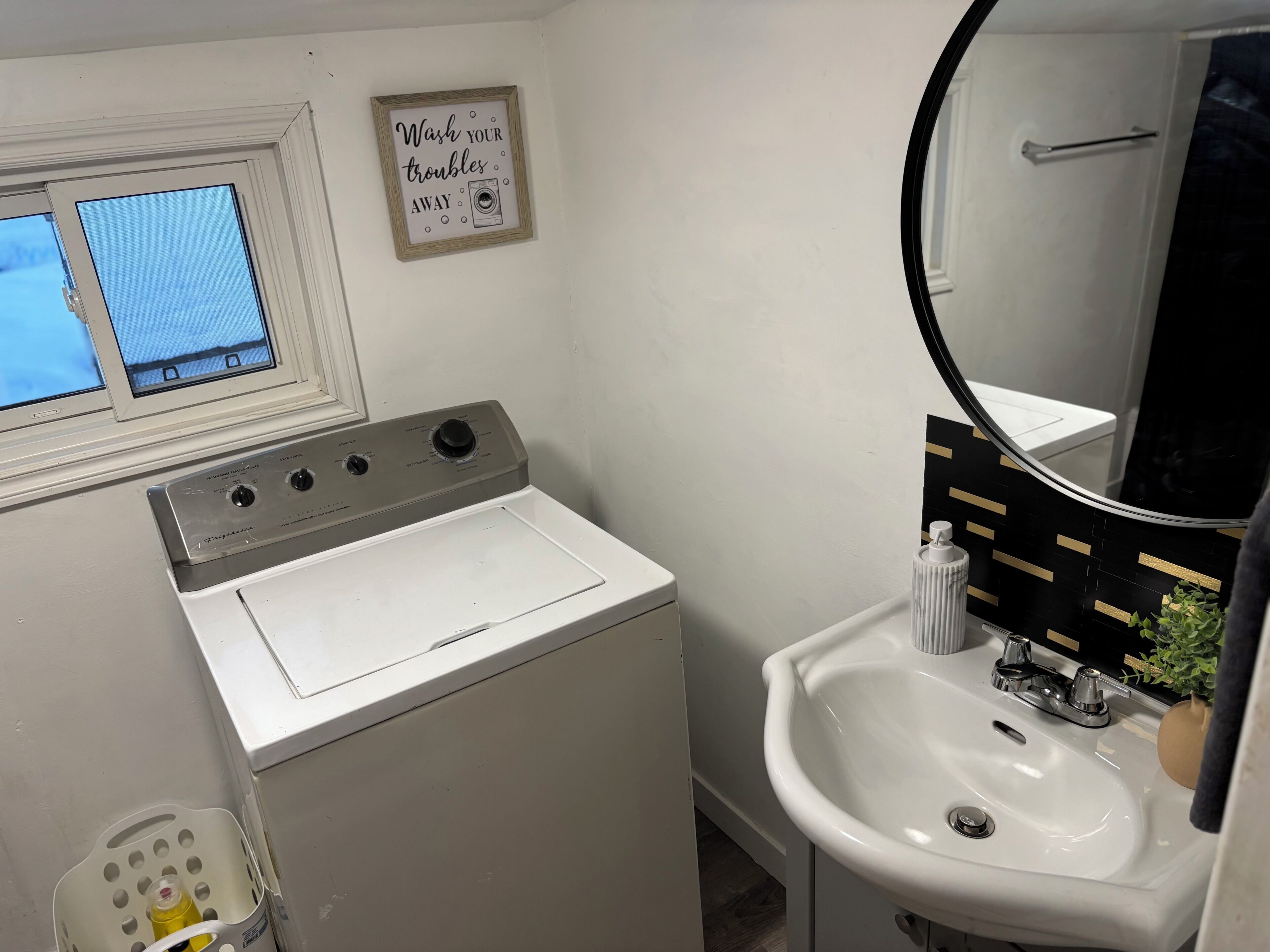 A compact bathroom with a washing machine, a sink, a mirror, and a window with a view.