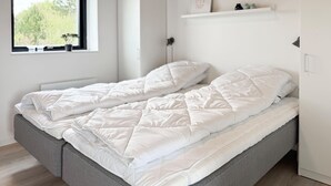 Una cama individual con estructura gris y ropa de cama blanca en una habitación con ventana, un armario blanco y un estante con objetos decorativos.